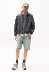 Giacca bomber grigia sopra un maglione blu a maglia, camicia gialla e shorts di denim chiaro. Cappellino grigio e sneakers bianche con accenti neri.