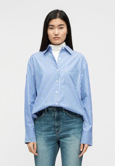 Νεαρή γυναίκα φορώντας μπλε και λευκό πουκάμισο με κάθετες ρίγες πάνω από λευκό ζιβάγκο, συνδυασμένο με μεσαίου τόνου μπλε τζιν, στέκεται μπροστά σε απλό φόντο.