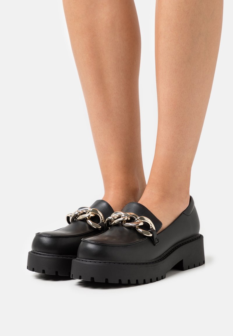 Chaussures style mocassin en cuir noir avec une semelle plateforme épaisse et un détail décoratif de chaîne argentée à l'avant. Texture lisse et forme arrondie.