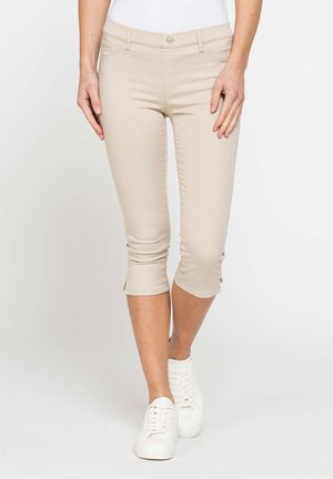 Pantaloni beige corti indossati con scarpe da ginnastica bianche e top bianco, mostrati dalla vita ai piedi su uno sfondo semplice.