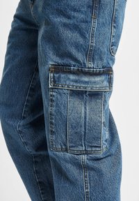Denim-Cargo-Hosen mit lockerer Passform, tiefen Seitentaschen, blassem blauen Farbton, robuster Nahtführung und leicht strukturiertem Stoff.