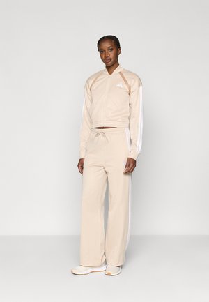 Beiger Trainingsanzug aus glattem Stoff mit weißen Seitenstreifen und gerippten Bündchen. Die Jacke hat einen Reißverschluss und ein Logo auf der Brust.