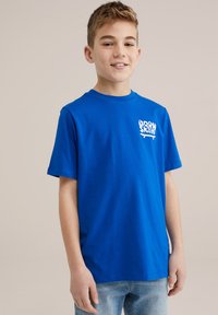 Blå bomullströja med korta ärmar, som har ett vitt grafiskt tryck med texten "BORN TO SKATE." Standard rund halsringning och avslappnad passform.