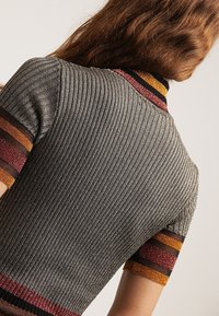 Haut à manches courtes en maille côtelée grise, avec des accents rayés colorés aux manches et au col en rouge, orange et noir.