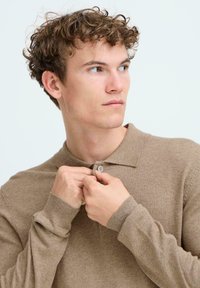 Camisa polo de punto marrón con cuello, mangas largas y un solo botón en el cuello. La textura parece suave y lisa.
