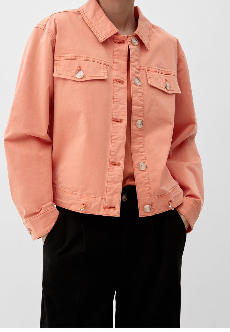 Personne portant une veste corail à boutons avec poches à rabat par-dessus un haut corail assorti et un pantalon noir, debout les mains dans les poches.