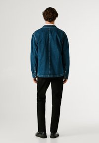 Giubbotto di denim blu scuro con vestibilità rilassata, polsini con bottoni e colletto classico, abbinato a pantaloni neri e calzature nere.