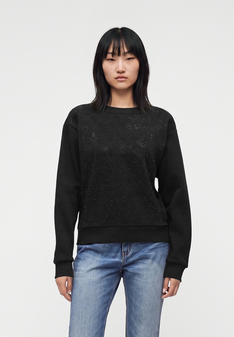 Jeune femme aux cheveux noirs et raides, portant un sweat-shirt noir à motifs et un jean bleu, debout devant un fond clair uni.