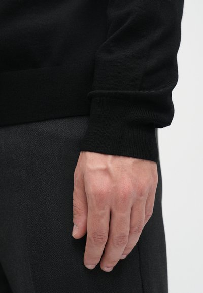 Pull noir avec des poignets côtelés porté sur un vêtement texturé gris. Gros plan d'une main reposant à côté d'un tissu foncé.