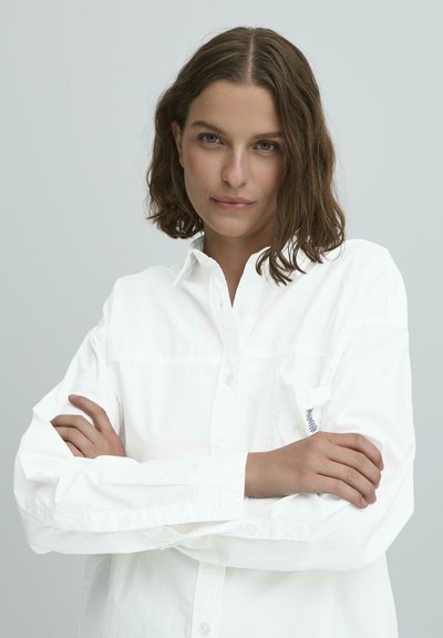 Balta ilga rankovė marškiniai, pagaminti iš medvilnės, su mygtukų užsegimu ir krūtinės kišene su kontrastiniu raštu. Lygus paviršius.