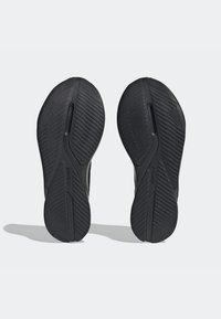 Une paire de semelles de chaussures de sport noires avec des motifs de rainures diagonales et un marquage "Adiwear", vues par le dessous sur un fond uni.