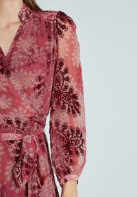 Wickelkleid aus weichem, transparentem, tiefrosa Stoff mit Blumenmuster, ausgestattet mit puffärmeligen langen Ärmeln und einem Bindegürtel in der Taille.