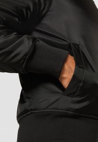 Chaqueta ligera negra con una textura suave, puños acanalados, bolsillos laterales y un diseño elegante. Presenta un brillo sutil y costuras reforzadas.