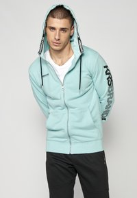 Sudadera con cremallera de color azul claro, con capucha forrada, cordones negros, bolsillos laterales y un logo en el pecho y la manga. Combinada con pantalones negros.