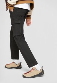 Black cargo pants with side pockets, paired with beige and black climbing shoes featuring red laces, and white ankle socks.
