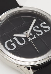 Reloj redondo negro con esfera texturizada, logo plateado "GUESS" y manecillas plateadas. Bezel metálico y correa negra.