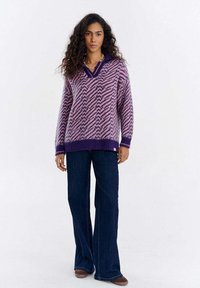 Kobieta o kręconych włosach, ubrana w fioletowy sweter w wzory, ciemne szerokie jeansy i brązowe buty, stojąca na jednolitym tle.