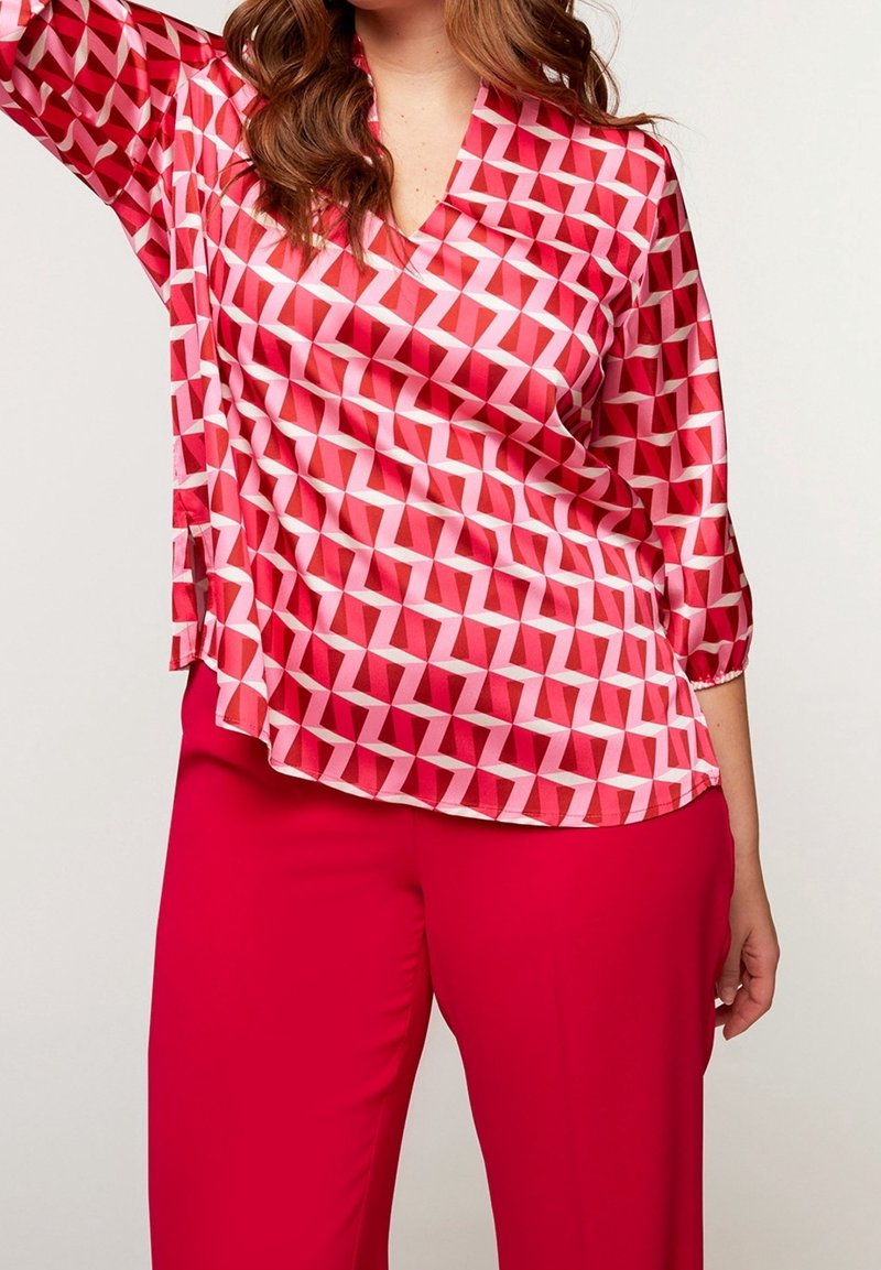 Femme portant un chemisier rouge à motif géométrique avec des manches trois-quarts et un pantalon rouge uni, posant avec un bras levé.