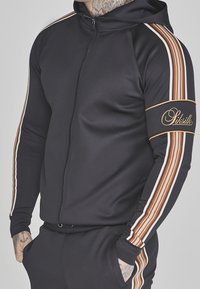 Sweat à capuche noir zippé avec une capuche, présentant des rayures verticales blanches et marron le long des manches et un logo de la marque sur le bras.