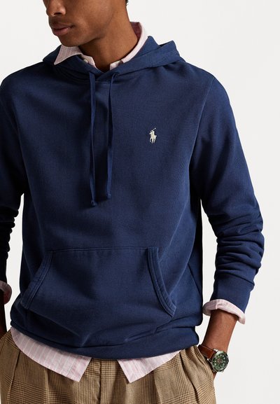 Tamnoprta kapuca iz teksturirane tkanine ima prednji žep i kapuljaču s vezicama. Izvezeni logotip je na prsima. Nosi se preko prugaste majice.