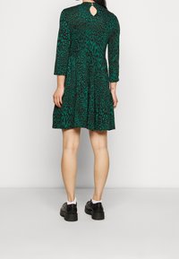 Grön klänning med leopardmönster, trekvartslånga ärmar, lager-på-lager kjol, nyckelhål i ryggen, bärs med svarta, kraftiga skor.