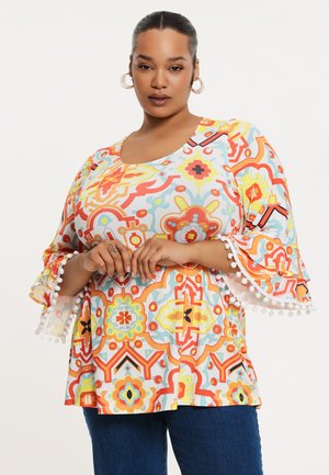 Bunt gemusterte Bluse mit Glockenärmeln und Pompon-Besatz, aus leichtem Stoff gefertigt. Das Design umfasst geometrische Formen in Orange, Blau und Gelb.