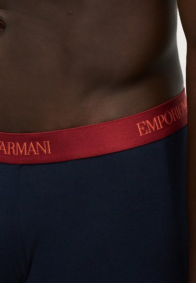 Gros plan sur la taille d'une personne portant un pantalon bleu marine avec une ceinture rouge montrant partiellement le logo Emporio Armani.