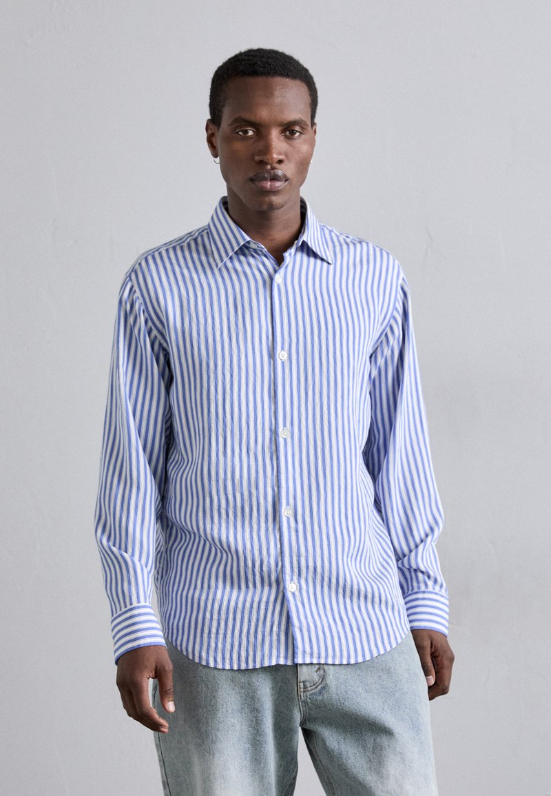Camisa a rayas azul y blanca con botones, de manga larga, cuello y un corte holgado, combinada con jeans azul claro. Textura de tela suave.