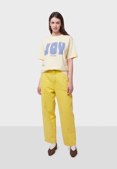Gule brede jeans og en lys gul T-shirt med blå "JOY" tekst. T-shirten har korte ærmer og en afslappet pasform. Brune sko.