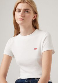 Tricou alb cu mânecă scurtă, fabricat dintr-un material cu structură ribbed, având un mic logo roșu Levi's pe partea stângă, asortat cu blugi bleu din denim.