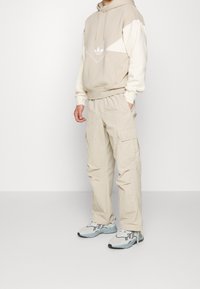 Felpa beige con dettagli bianchi e logo, abbinata a pantaloni cargo beige chiaro. Completa l'outfit un paio di sneaker azzurro chiaro con dettagli neri.