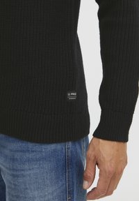 Fekete kötött pulóver bordás mandzsettával és aljjal, oldalán egy kis fehér címkével. Kék farmernadrággal viselve.