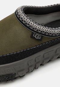 Zöld skór slip-on cipő fekete gumitalppal, texturált felülettel, kontrasztos varrással és "UGG" logós címkével. Díszes szegélyezéssel kiemelve.