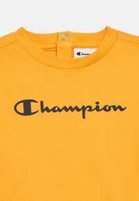 Κίτρινο βαμβακερό t-shirt με στρογγυλή λαιμόκοψη και κλείσιμο με κουμπί στο πίσω μέρος. Δια θέτει μαύρο λογότυπο "Champion" μπροστά.