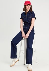 Combinaison en denim à manches courtes, fermeture éclair, et deux poches poitrine ; présente des coutures contrastées et un design à jambes évasées. Associée à une casquette rouge.