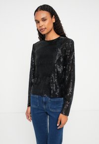 Femeie cu păr creț, îmbrăcată cu un top lung cu textură negru și blugi albaștri, stând în fața unui fundal alb simplu.