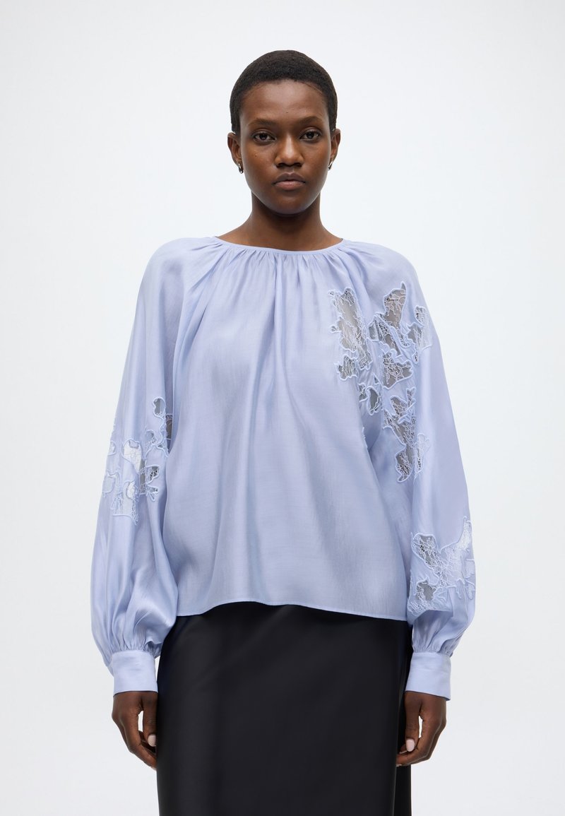 Γαλάζιο μπλουζάκι με μακριά φουσκωτά μανίκια, σούρες στο λαιμό και κεντήματα λουλουδιών με δαντέλα στα μανίκια. Φορεμένο πάνω από μαύρη φούστα.