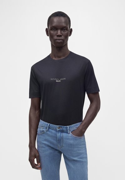 Άνδρας με σκούρο δέρμα που φοράει μαύρο T-shirt Emporio Armani Milano και ανοιχτό μπλε τζιν σε απλό λευκό φόντο.