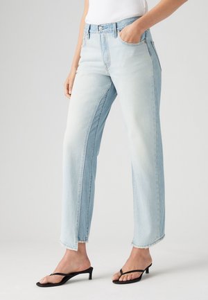 Persoon die lichtgewassen jeans met rechte pijpen en onafgewerkte zoom draagt, een wit shirt en zwarte sandaaltjes met kittenhiel, staand met één hand in de zak.