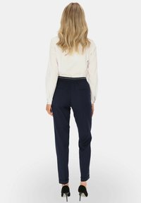 Kobieta z falującymi blond włosami, ubrana w białą bluzkę z długim rękawem, granatowe spodnie skrojone na miarę z mankietami oraz czarne szpilki, stoi tyłem.