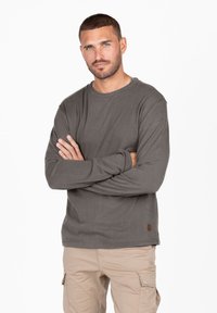 Graues Langarmshirt aus weicher Baumwolle, mit Rundhalsausschnitt und dezentem Logo-Detail am Saum. Getragen mit beigen Hosen.