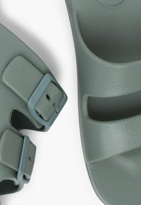 Sandales en plastique vert avec des sangles réglables, présentant une surface texturée et deux boucles latérales pour un maintien sûr.