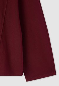 Tessuto bordeaux con una texture liscia e cuciture visibili. Presenta un design morbido e drappeggiato con una leggera svasatura alle maniche.