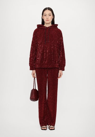 Red sequin hoodie with drawstring and matching pants. Textured, loose fit, paired with black strappy sandals and a woven handbag.
