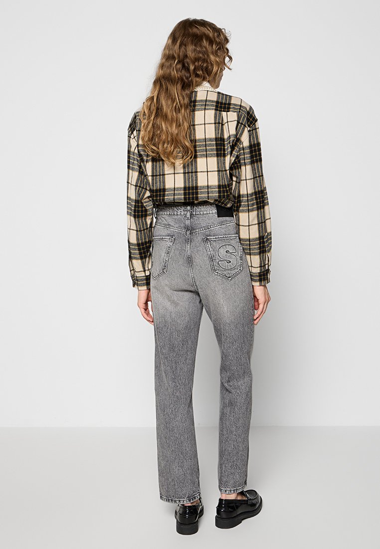 Koszula w kratę w kolorze beżowym i czarnym, zestawiona z wysokim stanem szarych dżinsów z logo z tyłu i prostymi nogawkami. Czarne buty.