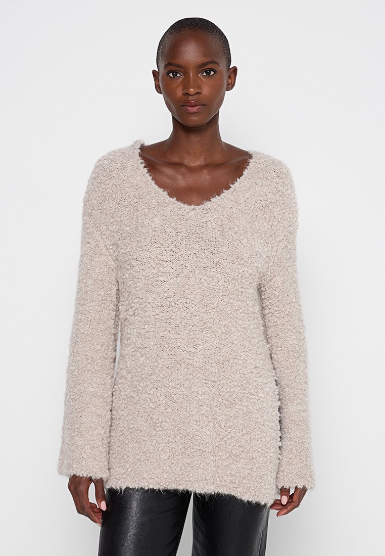 Beige gebreid trui met een diepe V-hals, lange mouwen en een gestructureerde afwerking, met een losse pasvorm en rafelige zomen.
