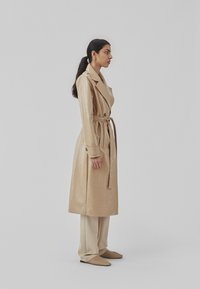 Beige læder trench coat med lange ærmer, bælte i taljen, store revers og shiny tekstur, stylet med cremede bukser og vævede slip-ons.