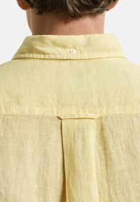 Gelbes Leinenhemd mit einem knöpfbaren Kragen und Rückenfalten, das eine weiche Struktur und ein leichtes Material zeigt.