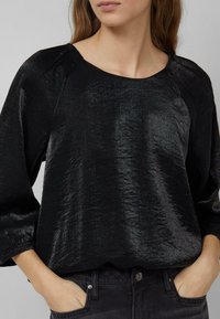 Schwarzes Oberteil aus glänzendem, strukturiertem Stoff mit rundem Ausschnitt, Dreiviertelärmeln und gesammelten Bündchen. Kombiniert mit dunkler Jeans.
