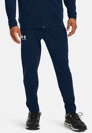 Mann trägt marineblaue Under Armour Trainingshose und passende Jacke, steht mit den Händen an den Seiten und schwarzen Laufschuhen.